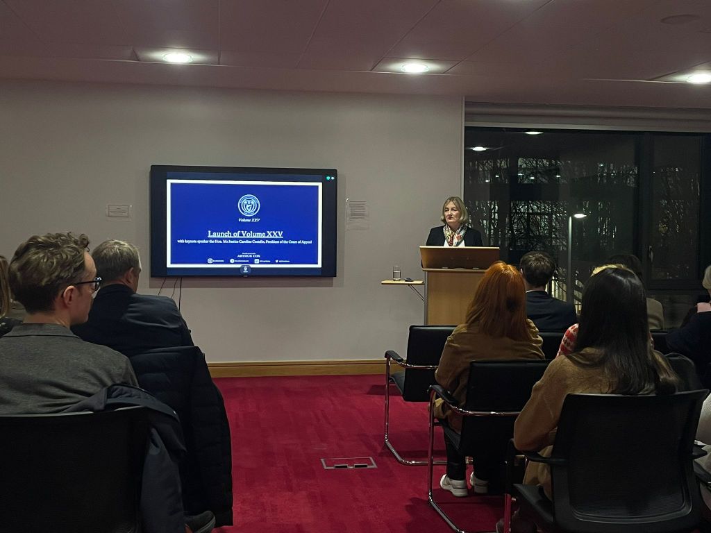 A speaker presenting at the launch event of the UCD Law Review Volume XXV, with an audience seated in front of a screen displaying the event details.